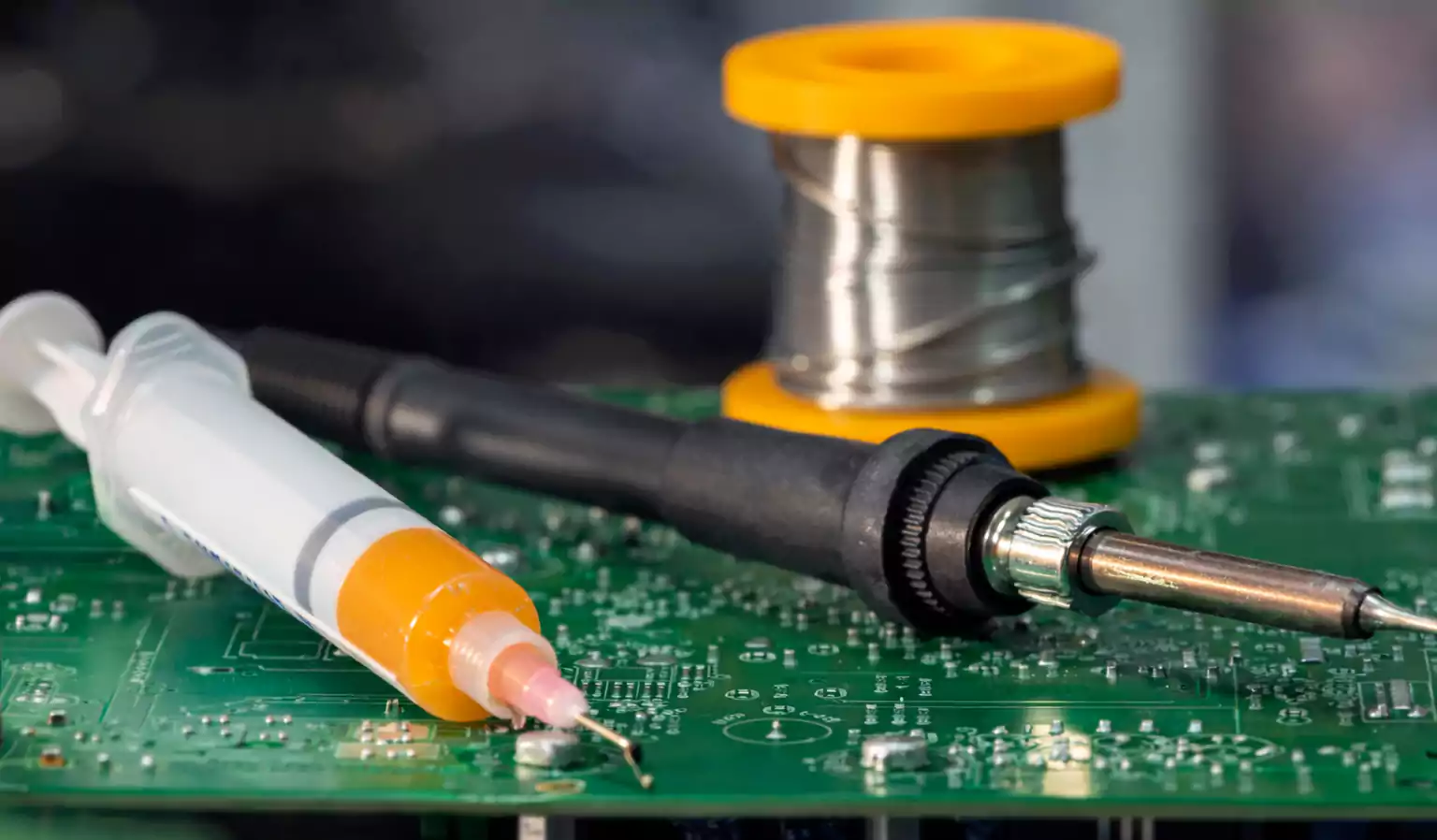What Is Flux in Soldering: soldering iron, flux syringe, and solder wire on a green PCB.